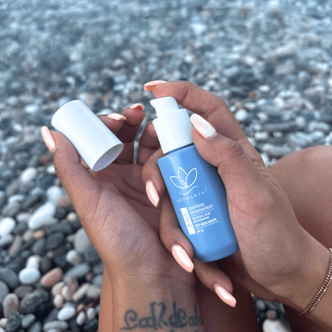 A backdrop of the Greek pebble beach with a woman's hands holding the bottle with cap open about to apply moisturizer