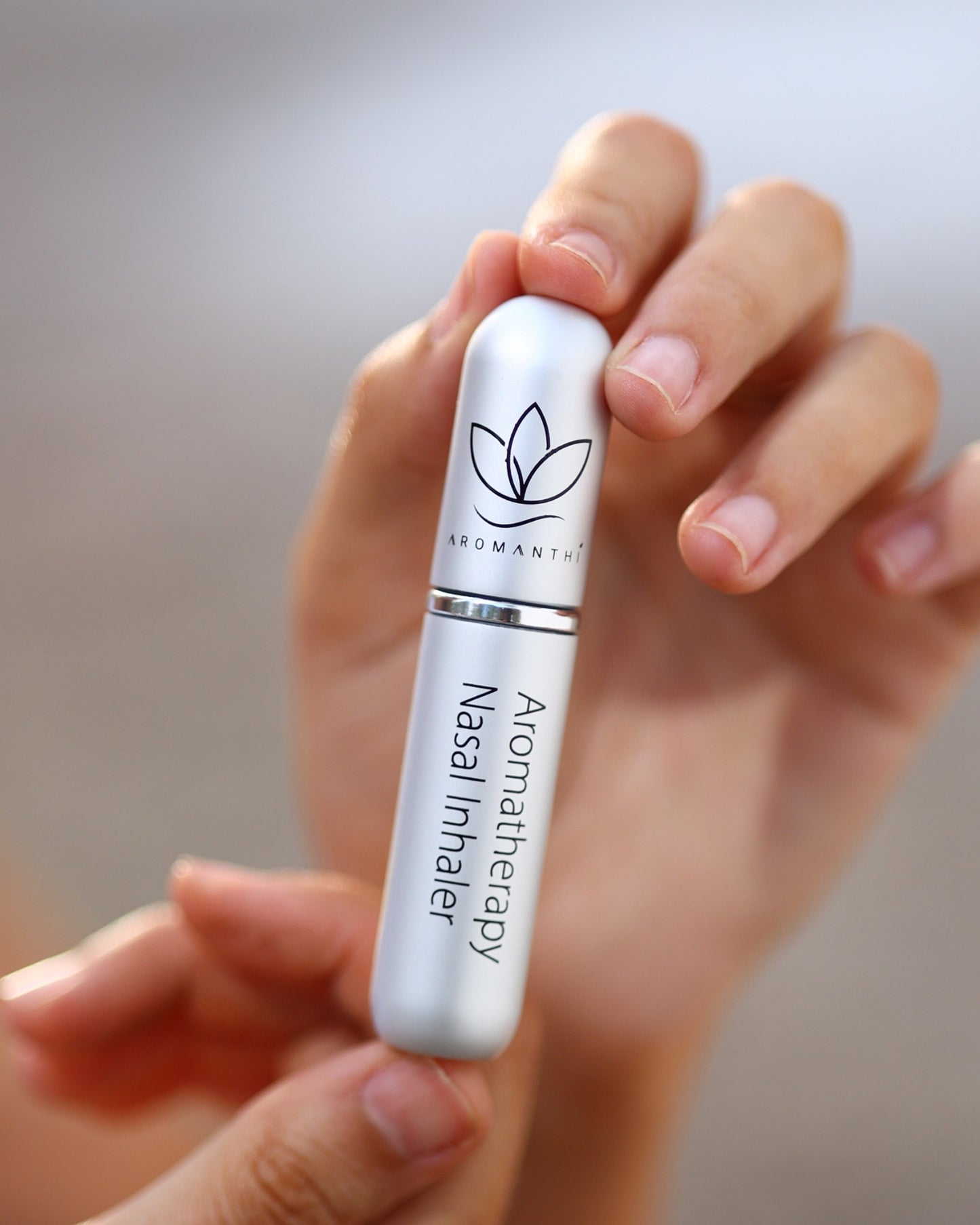 Aromanthi's Aromatherapy nasal inhaler held in a hand with a blurred background. Inhaler is the silver color option