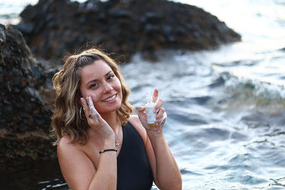 a woman with the background of the ocean holding aromanthi facial cleanser and applying it to her cheek.